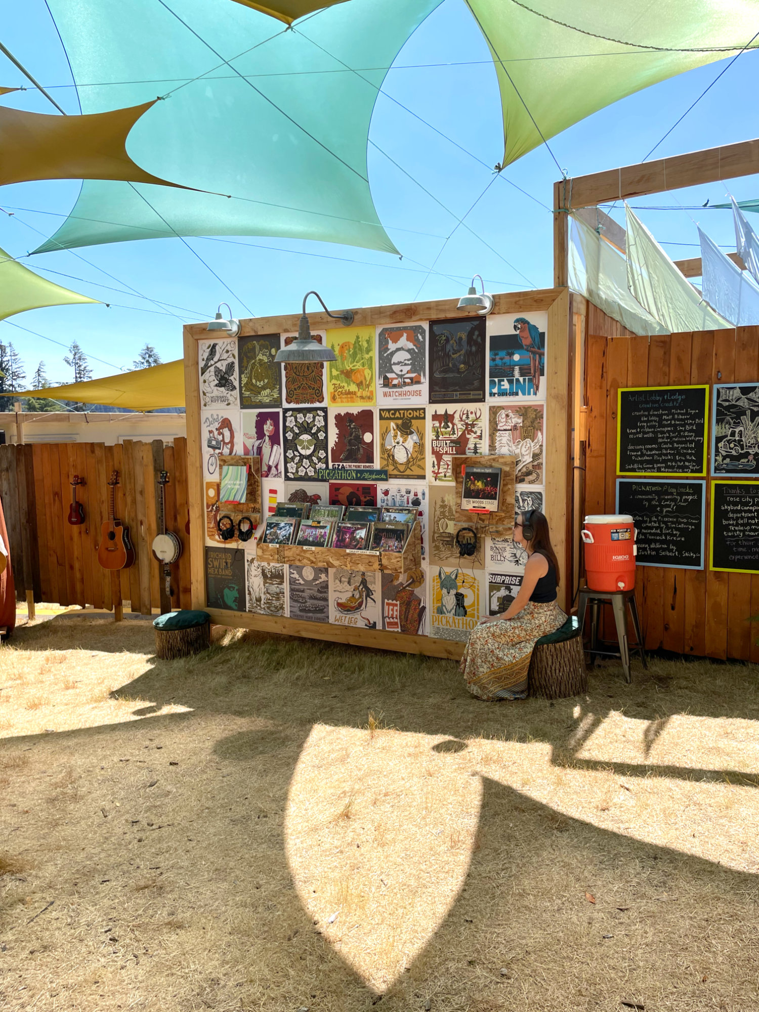 A wide view of the Pickathon Playbacks installation in the 2026 Artist Lodge: the poster-covered crate wall, two listening stations, and a visitor seated with headphones beneath the shade sails.
