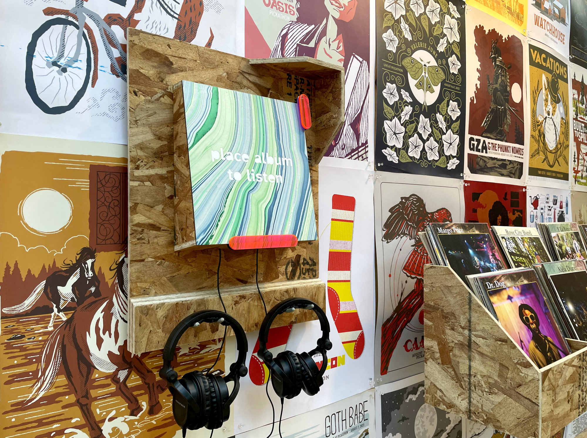 Close-up of a hand-built listening station: a teal and green marbled surface on a wooden shelf labeled “place album to listen,” with headphones hanging beneath.