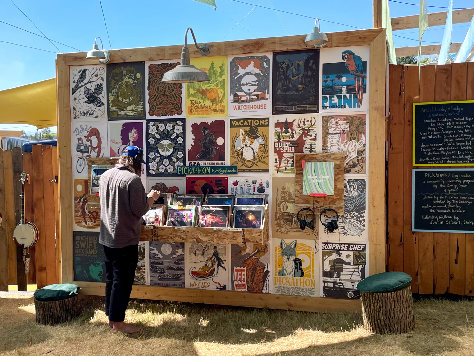 A visitor flips through record-style albums in a wooden crate mounted on a wall covered in Pickathon posters, with headphones and a listening station reading “place album to listen” beside it.